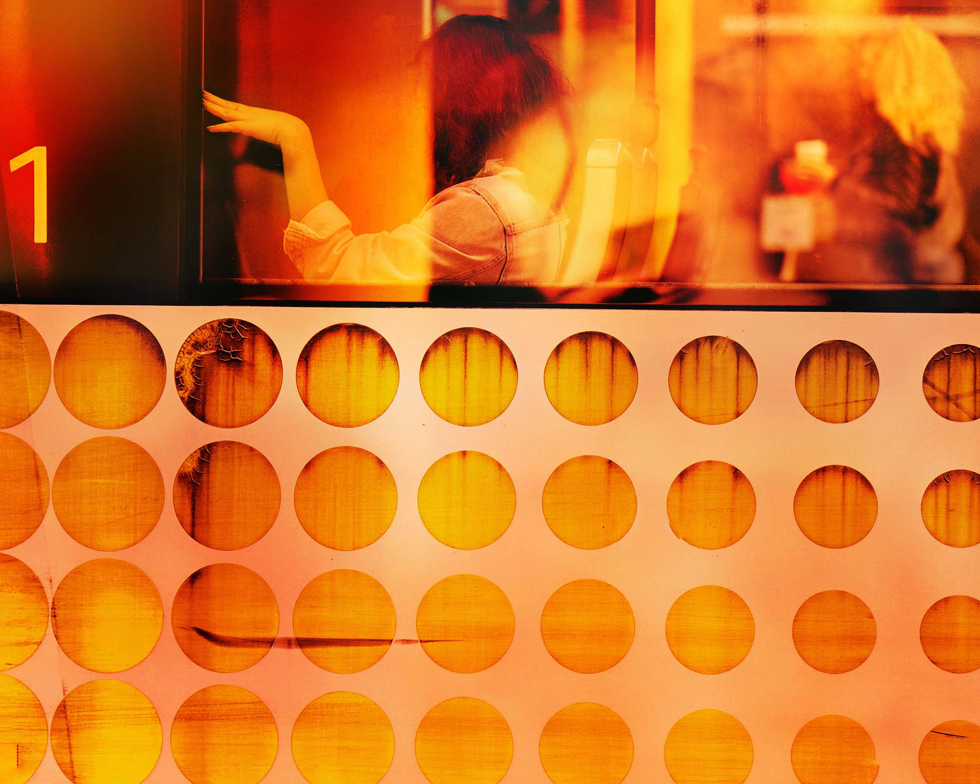 http://A%20warm-toned,%20blurry%20image%20from%20inside%20a%20vehicle,%20likely%20a%20bus%20seen%20through%20a%20window%20and%20panel.%20The%20upper%20window%20shows%20a%20person%20with%20reddish%20hair%20from%20behind,%20their%20hand%20extended,%20and%20the%20number%201%20on%20the%20left.%20The%20lower%20panel%20has%20rows%20of%20circular%20openings%20revealing%20a%20textured%20orange%20surface.