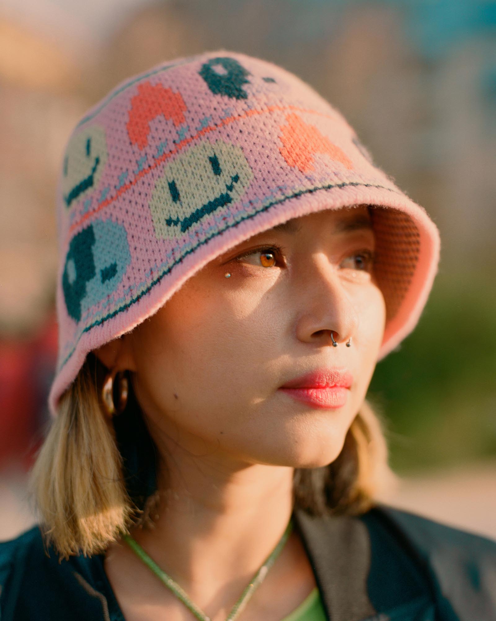 http://Close-up%20of%20a%20person%20wearing%20a%20pink%20knitted%20bucket%20hat%20with%20smiley%20faces,%20looking%20off%20to%20the%20side%20in%20warm%20sunlight