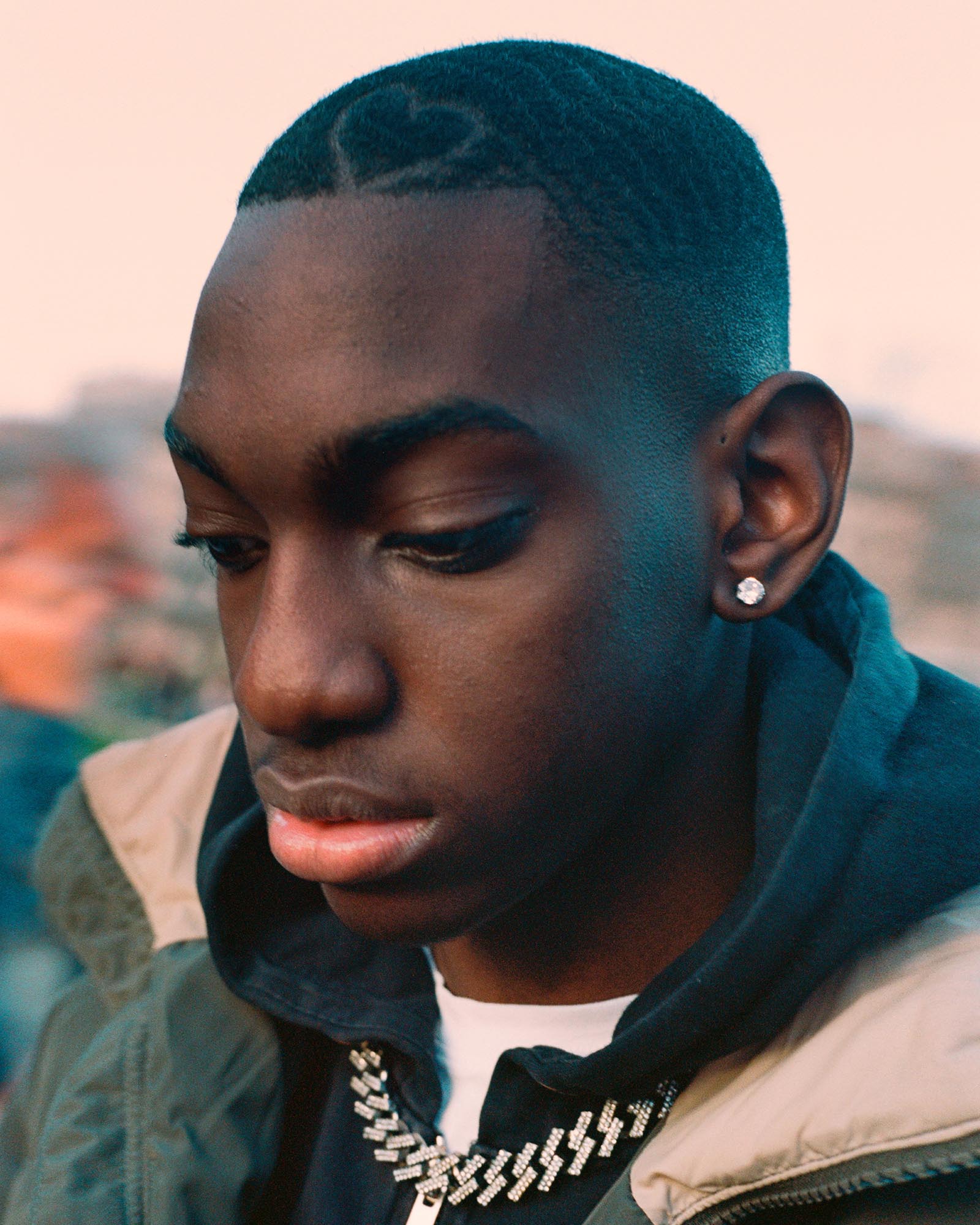 http://Close-up%20of%20a%20young%20man%20looking%20down,%20with%20a%20heart%20shape%20shaved%20into%20his%20short%20hair,%20wearing%20an%20iced-out%20chain%20and%20stud%20earring.