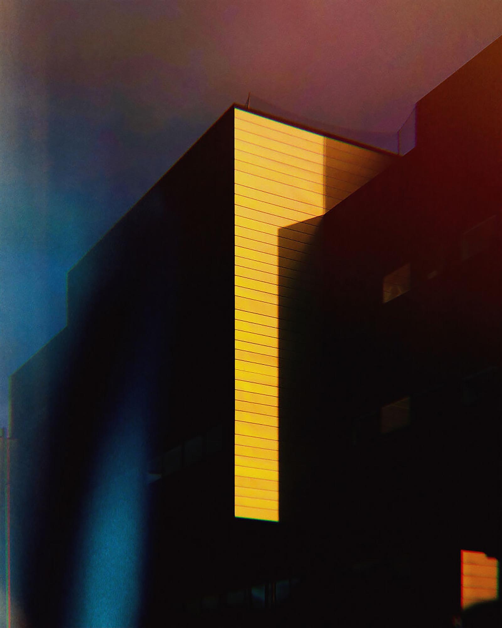 http://Abstract%20architectural%20detail%20of%20a%20building%20with%20a%20bright%20yellow%20vertical%20section%20against%20dark%20shadowed%20areas%20and%20a%20moody%20blue/red%20sky